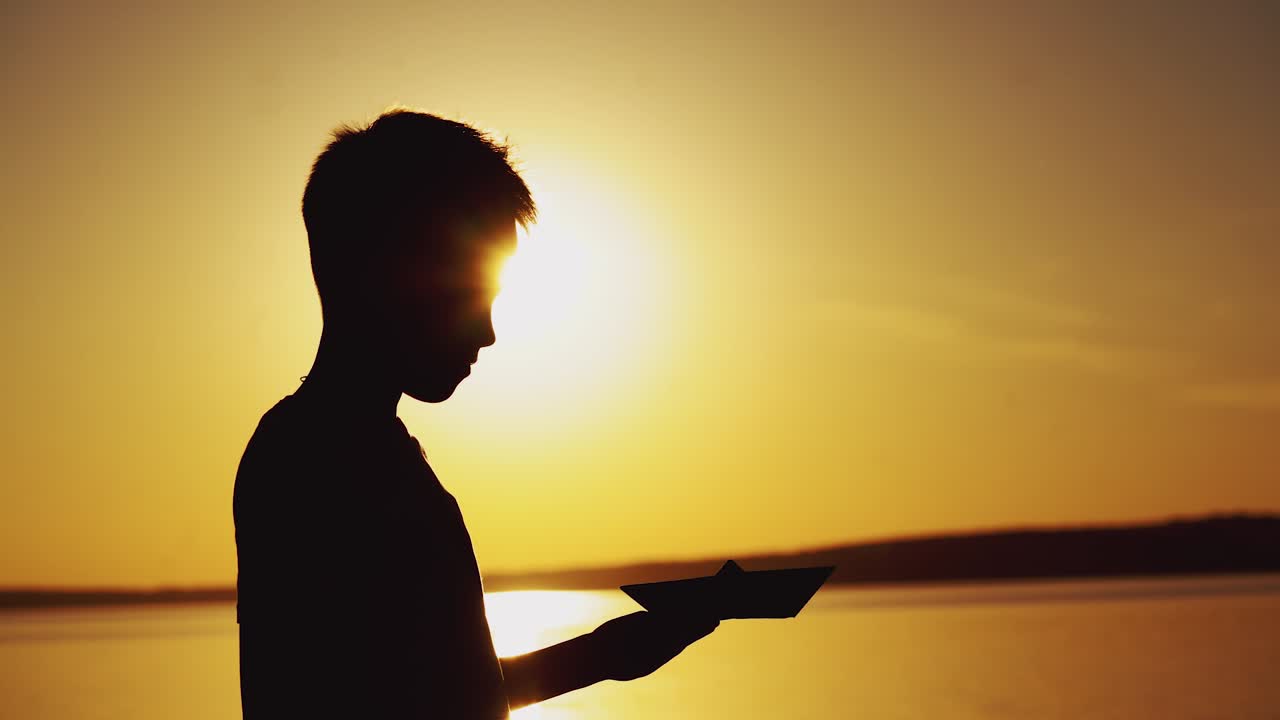 Silhouette di un ragazzo in piedi vicino al fiume che gioca con una barchetta di origami a un bellissimo tramonto. La barchetta di carta è nelle mani di un bambino piccolo e il sole splendente della sera