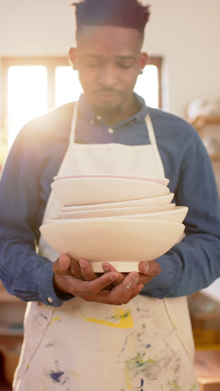 Happy african american potter holding white bowls and smiling in pottery studio, slow motion
