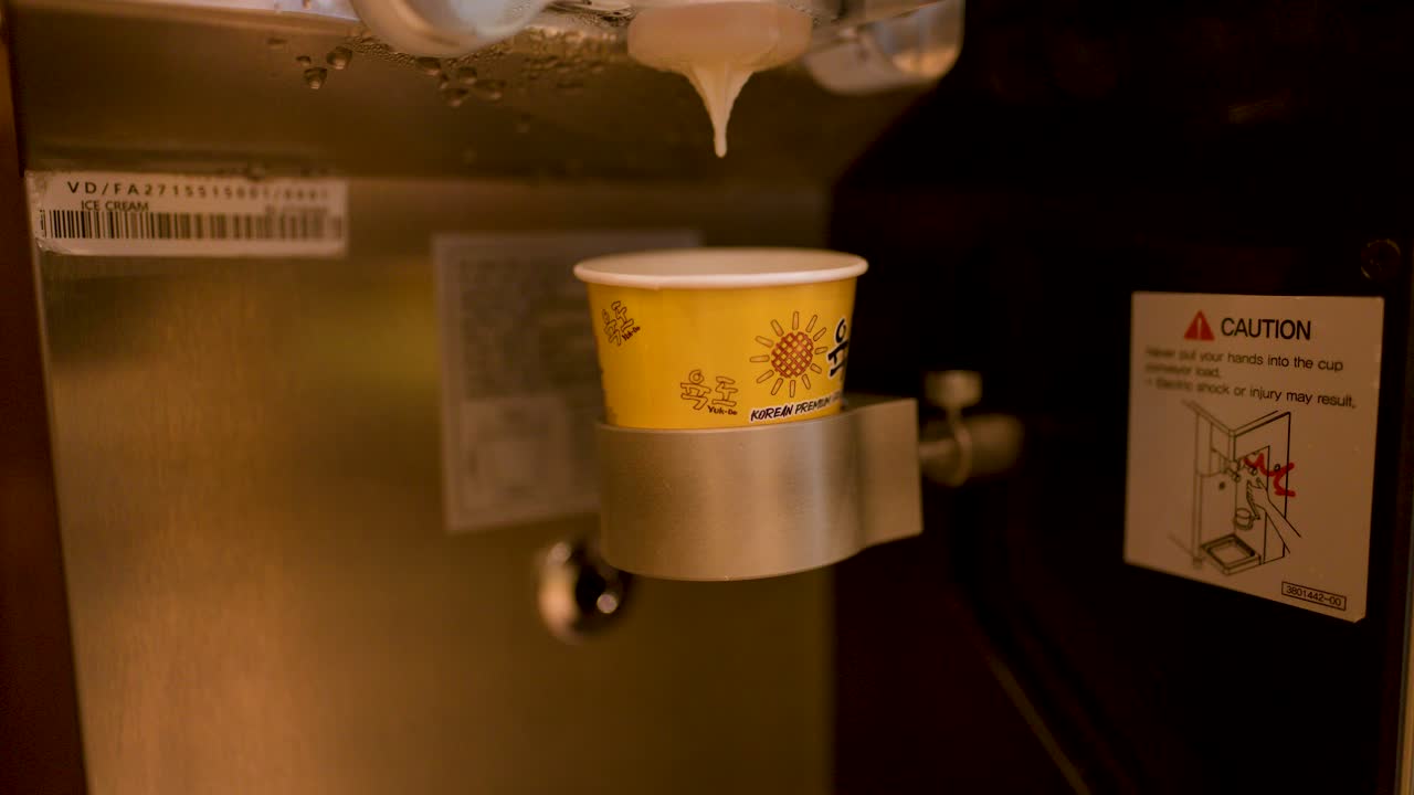 Hand positions paper cup under industrial dispenser, preparing for automated dessert serving, warm lighting