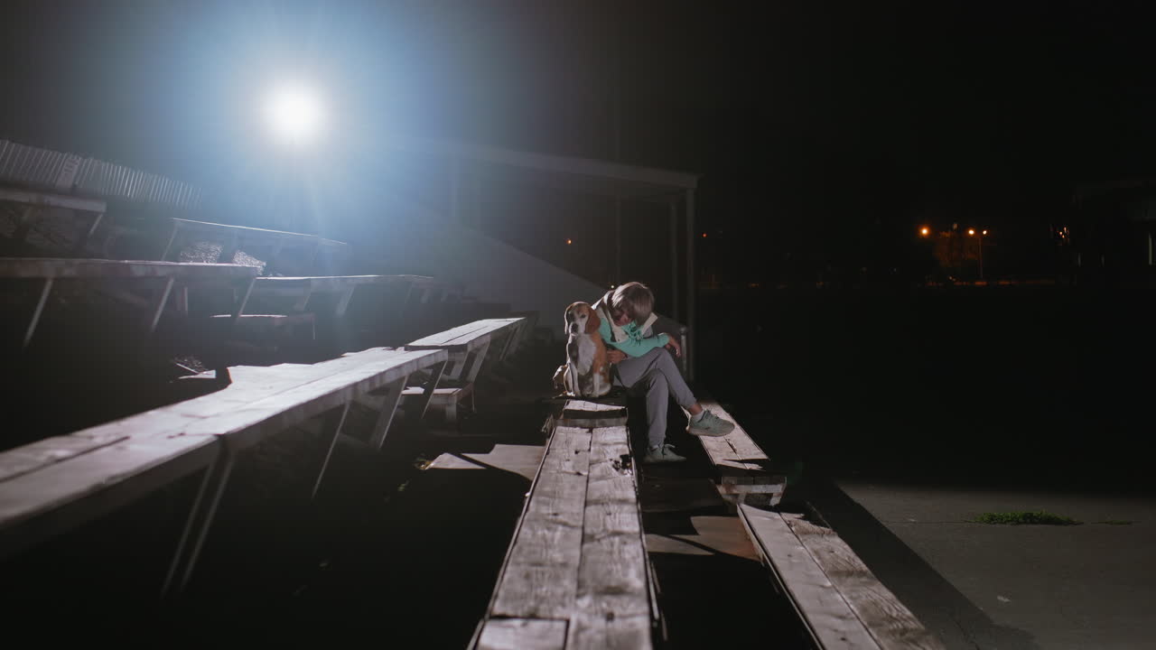 Trainer sitting quietly with beagle dog on wooden benches under bright spotlight at night after training session, sharing peaceful resting moment together in calm outdoor environment