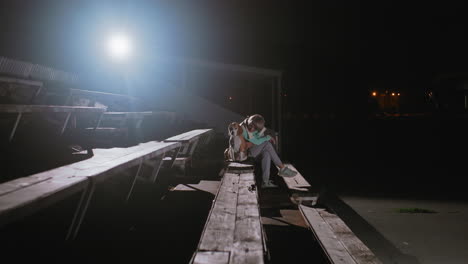 trainer sitting quietly with beagle dog on wooden benches under bright spotlight at night after training session, sharing peaceful resting moment together in calm outdoor environment