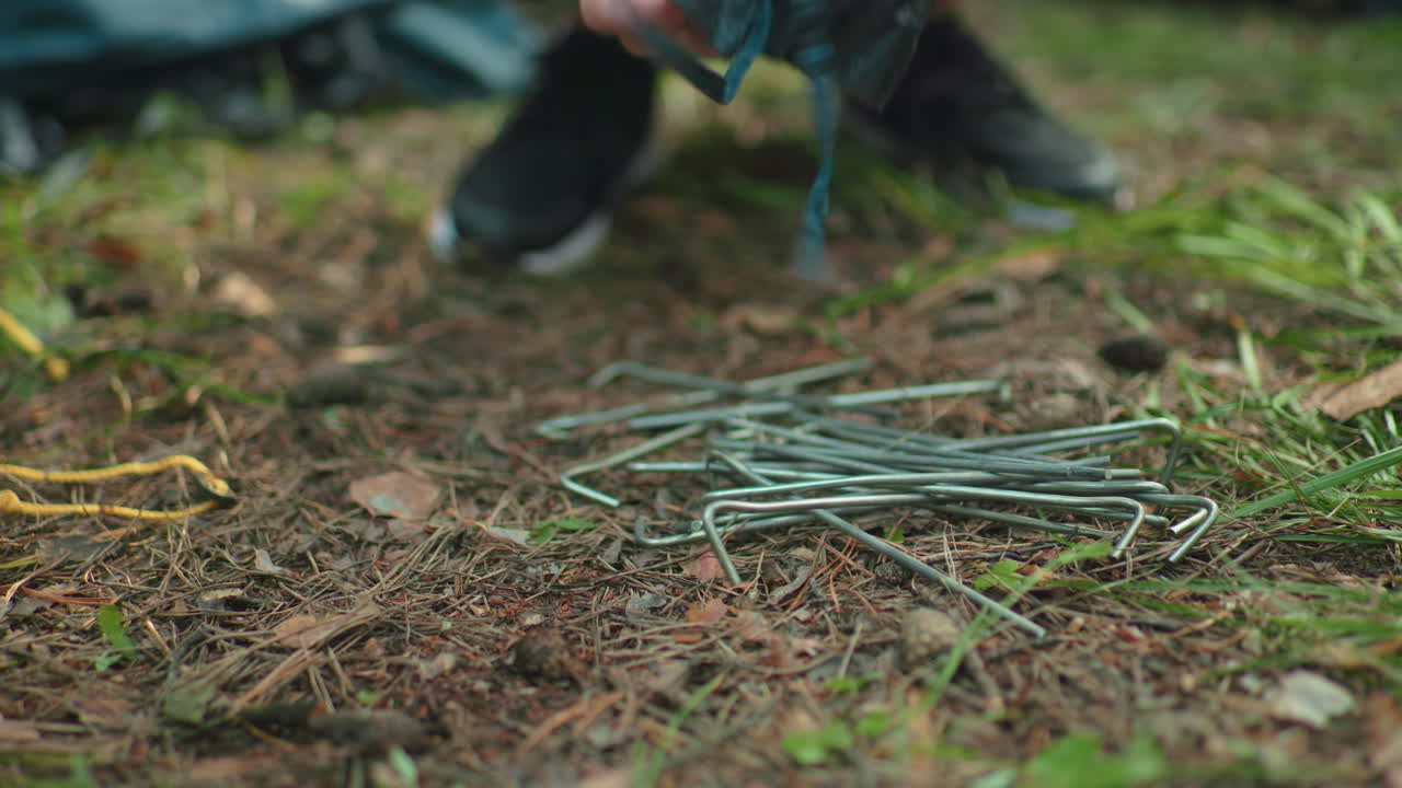 close up of hand turning camping equipment on forest ground while reaching for metal tent stakes among pine needles and grass with blurred shoes in background
