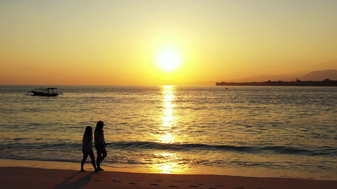 Two girls walking barefoot on sandy exotic beach washed by sea flows reflecting beautiful sunset with golden sky in Thailand