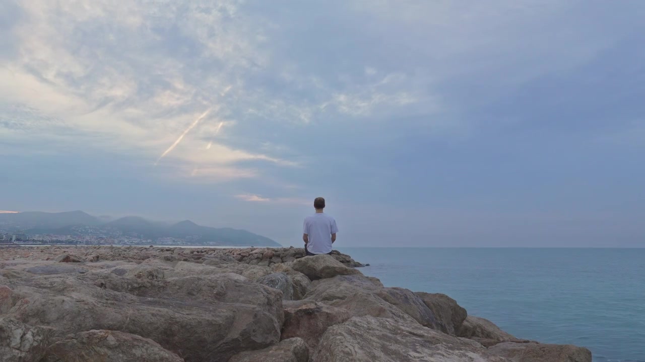 Il timelapse cattura un uomo solitario che medita sulle rocce in riva al mare mentre le nuvole sfrecciano in una giornata grigia