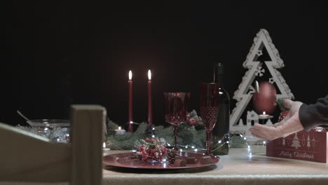 Man-takes-a-red-Christmas-tree-ball-on-a-decorated-table-in-a-dark-room-with-a-chair-in-foreground