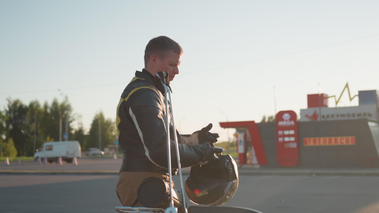 Young man in leather jacket and gloves takes helmet from motorcycle backrest and places it on seat while preparing to wear it, standing outdoors on city street under evening sunlight