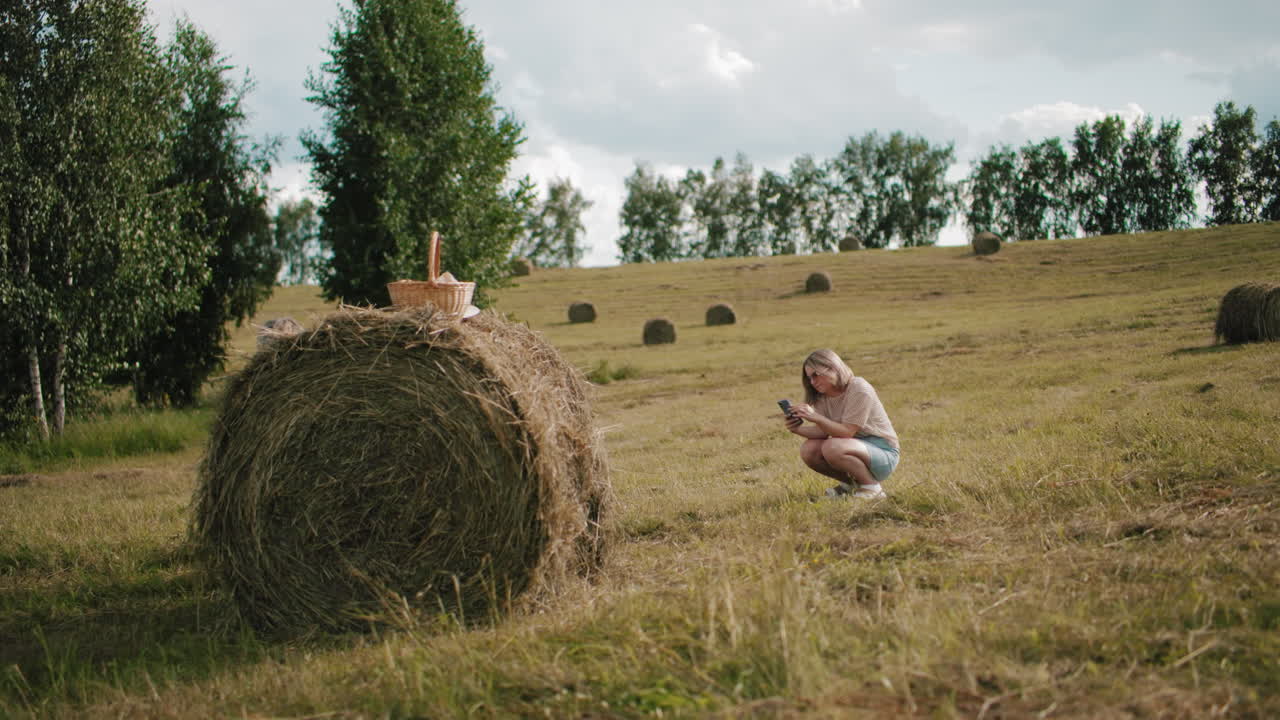 mujer agachada en un campo de hierba tomando una foto de una canasta de picnic en una bola de heno mientras otra mujer emerge de detrás, colinas rodantes y balas de heno esparcidas