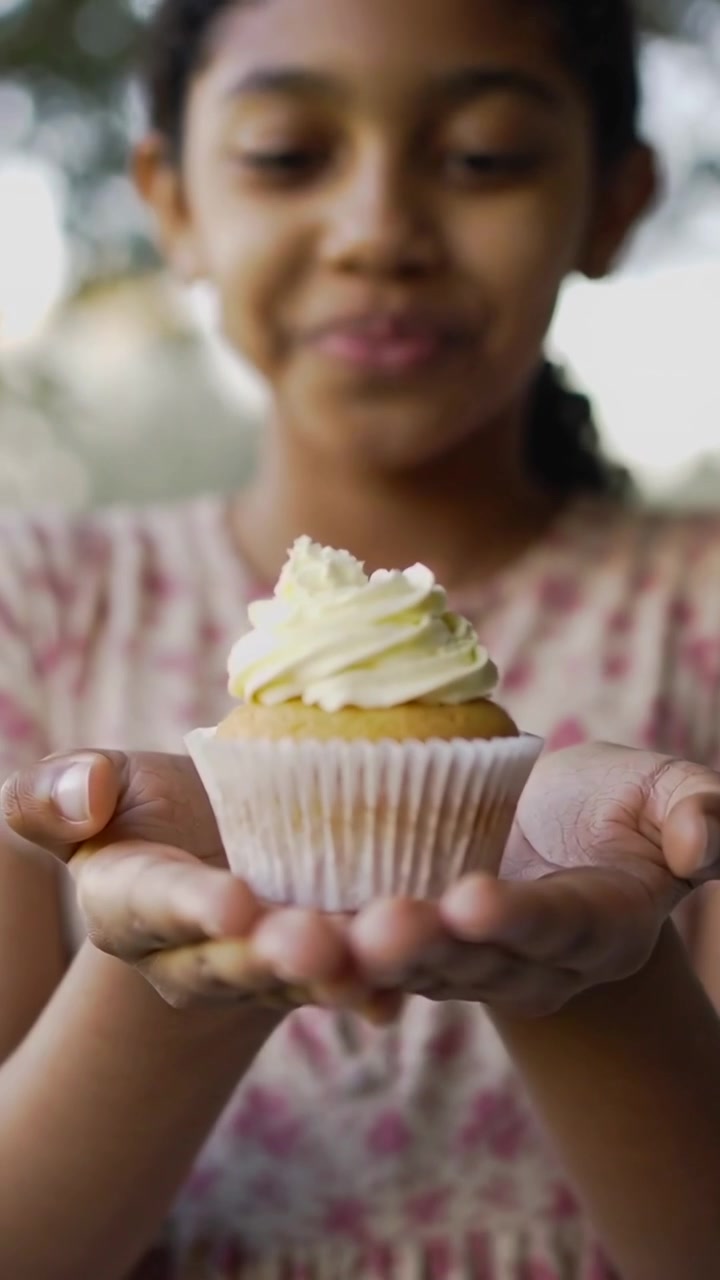 Video verticale: Ragazza con vestito floreale rosa in veranda che increspa le labbra, soffia e poi offre un cupcake.