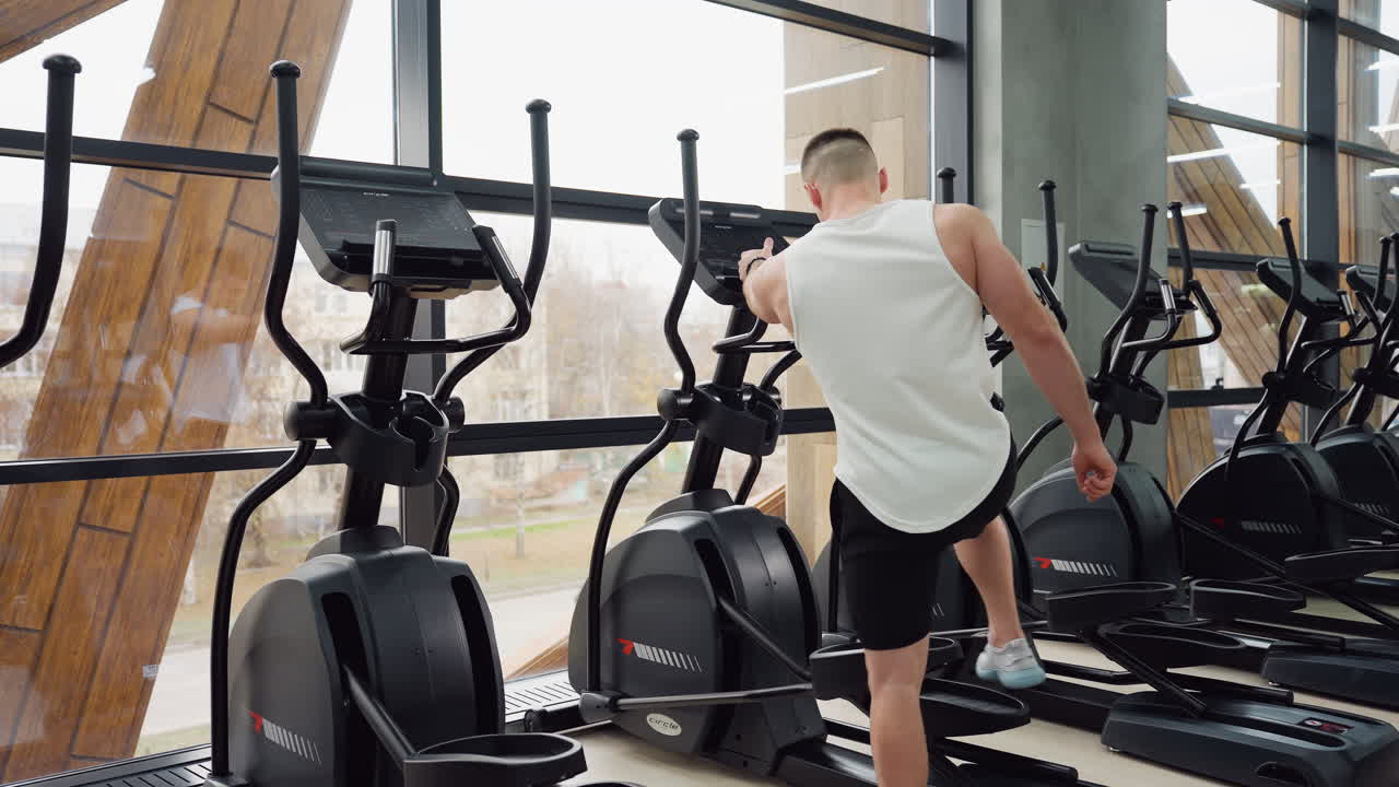 Man in white tank top black shorts walks toward elliptical trainer, stepping onto pedal in bright modern gym under large windows with urban view ready for cardio workout session focusing posture
