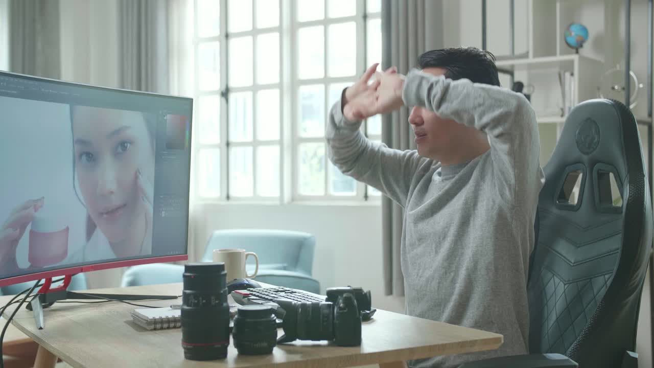Asian Cameraman In Long Sleeved T-Shirt Stretching And Leaning Back While Using Desktop Computer For Editing Photo At Home.