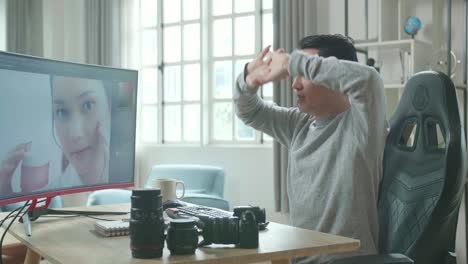 asian cameraman in long sleeved t-shirt stretching and leaning back while using desktop computer for editing photo at home.