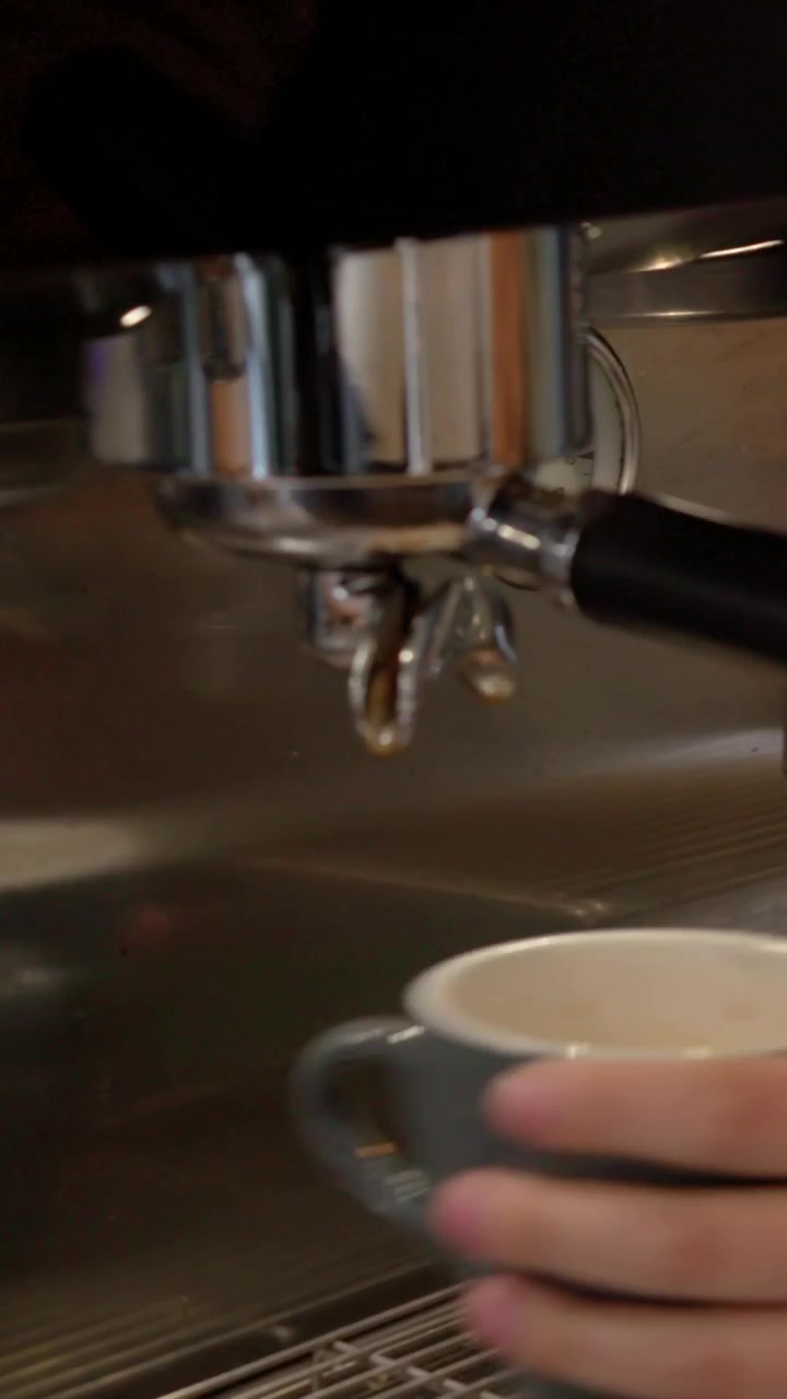 Video verticale: Avviso d'ordine, barista con grembiule verde che monta il latte in una lattiera e prepara un caffè espresso al bancone.