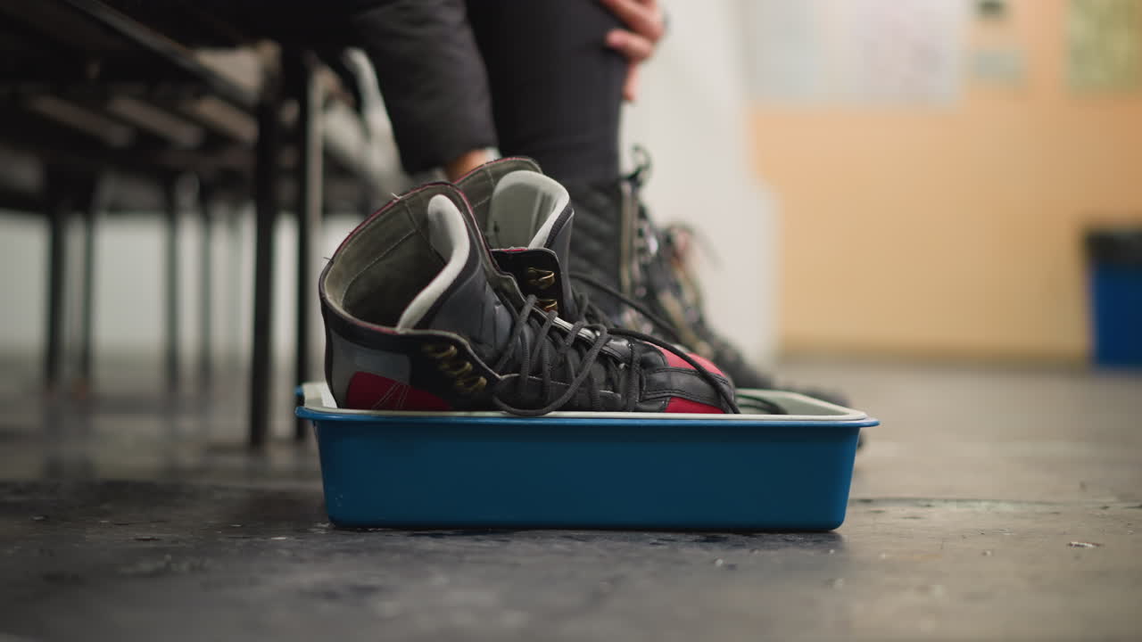 Hand changing shoe with black leather boot on scratched indoor floor holding blue plastic tray containing red and black ice skates preparation for skating activity visible bracelets and watch lifestyle