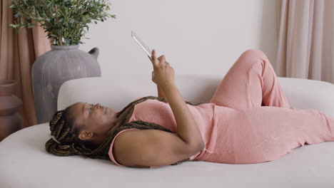 Woman-With-Braids-Lying-On-A-Big-Sofa-While-Using-A-Tablet