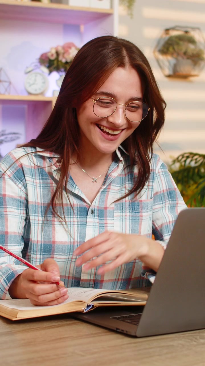 Mujer joven en la mesa de casa saludando a la cámara del portátil con gafas y libro para un seminario web o lección de estudio en línea