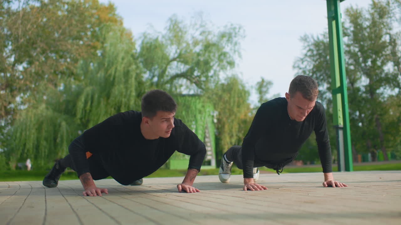 Due uomini che fanno flessioni all'aperto su una piattaforma di legno in un parco verde, partner di allenamento che eseguono ripetizioni con espressioni concentrate, abbigliamento sportivo, lavoro di squadra di supporto e respirazione costante