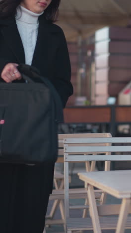 young investor in black coat relaxes at a cafeteria, setting her bag on the table against a blurred background with urban elements like poles, flowers, and buildings