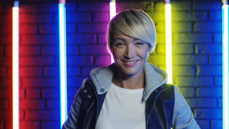 blonde woman with short hair standing on wall with neon lamps smiling and looking at camera