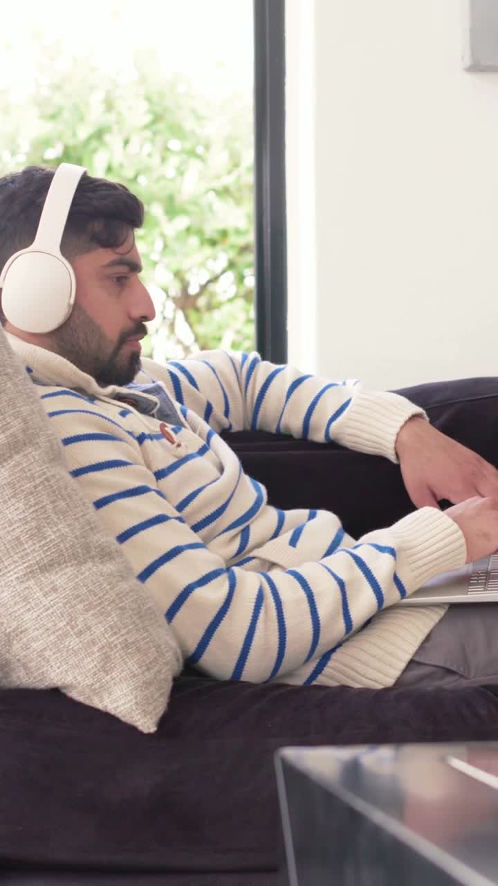 Video verticale: Uomo sdraiato con maglione a righe e cuffie che lavora al laptop vicino alla finestra
