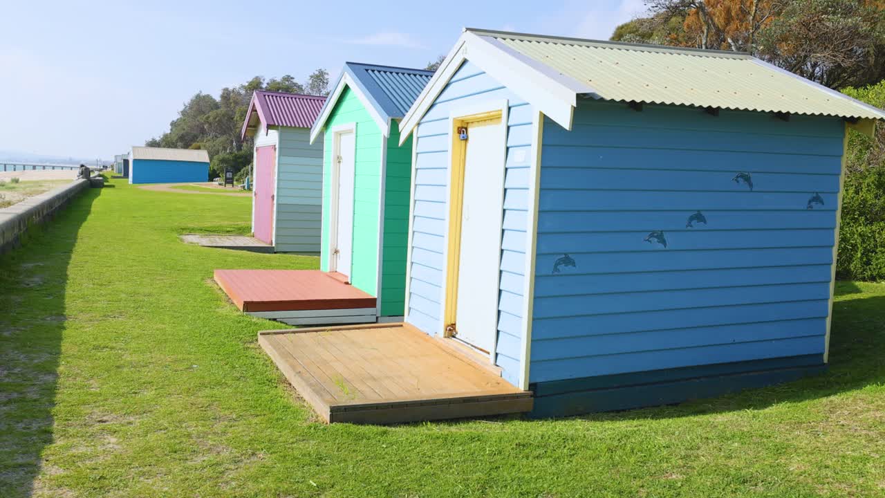 Vibrant beach huts line a grassy path under bright daylight, showcasing a serene coastal atmosphere