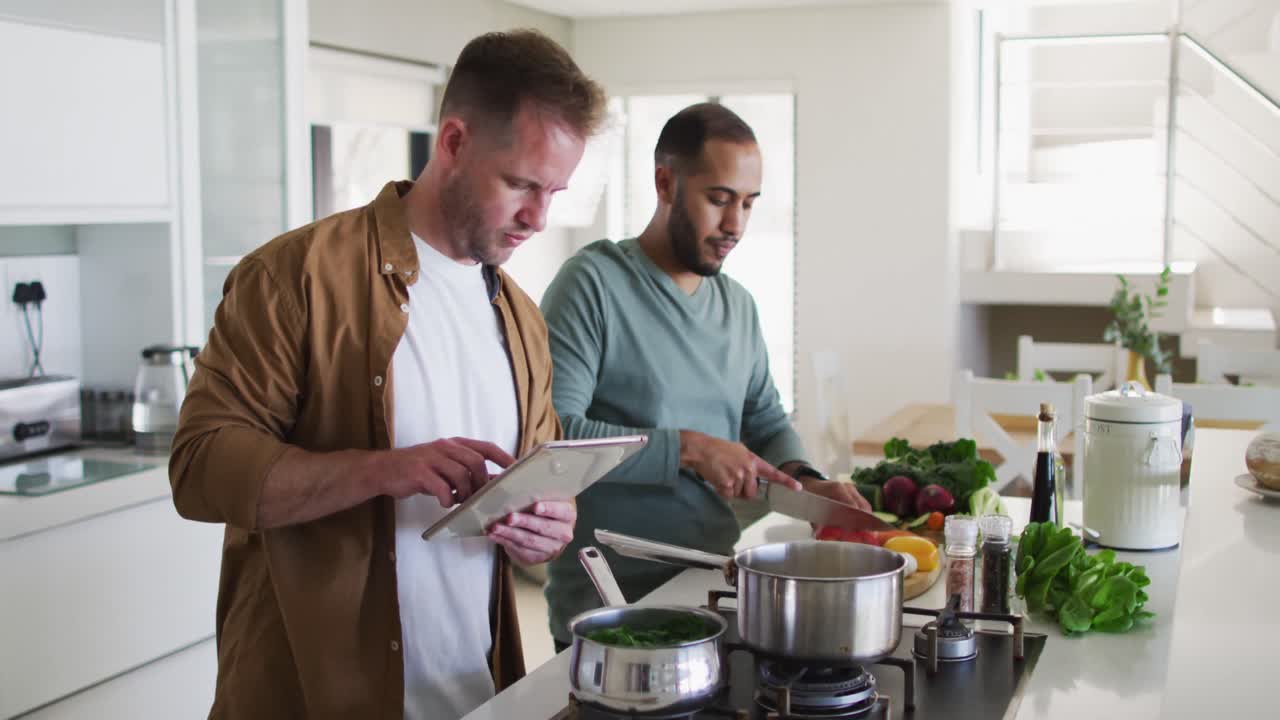 pareja de hombres homosexuales multiétnicos preparando comida en la cocina uno usando tableta