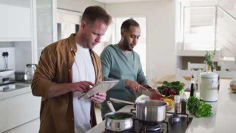 Multi-ethnic-gay-male-couple-preparing-food-in-kitchen-one-using-tablet