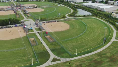 Seitliche-Und-Umlaufende-Luftaufnahme-Des-Ballparkkomplexes-Der-Vorstadtgemeinde-Mit-Mehreren-Laufenden-Softballspielen