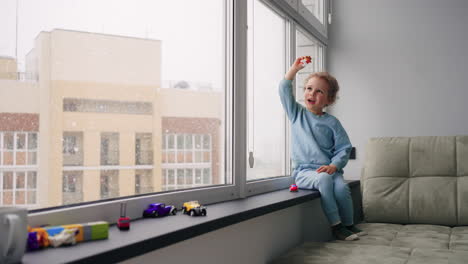 cute little boy is playing alone in children room in modern apartment in winter day snow is falling behind indow