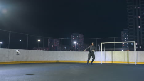 jogador de futebol em roupas esportivas escuras chuta a bola de futeblol para fora da área de gol em um campo urbano à noite, cercado por edifícios altos, esta ação dinâmica transmite determinação sob holofotes brilhantes