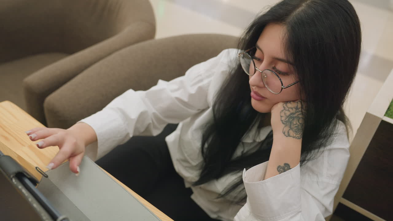 Top down view of lady in glasses working on tablet with one hand on screen and other resting under chin, floral tattoo visible on wrist, seated in soft chair with light wood table in indoor setting