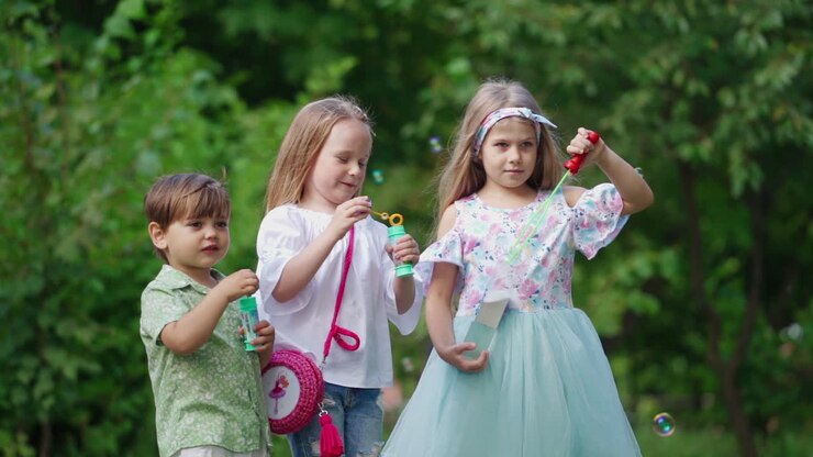 Happy kids have a rest in the park in a summer day. Little children blow air bubbles on the background of green bushes outdoors.