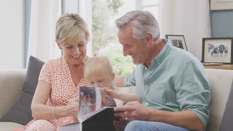 Abuelos-Sentados-En-El-Sofá-Con-Su-Nieta-En-Casa-Mirando-Juntos-Un-álbum-De-Fotos