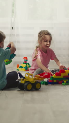 toddler boy and preschooler girl build house from colorful plastic blocks sitting on wooden floor. children enjoy playing with constructor at home