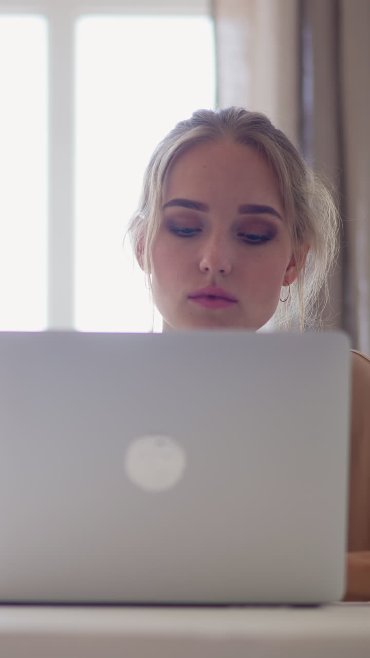 Young freelancer woman with fair hair works on modern laptop at white table with cup of hot drink in home office room against bright window slow motion