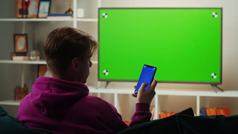 man watching television in living room. young guy sitting on sofa against tv with green screen, back view. male person using smartphone with blue chroma key.