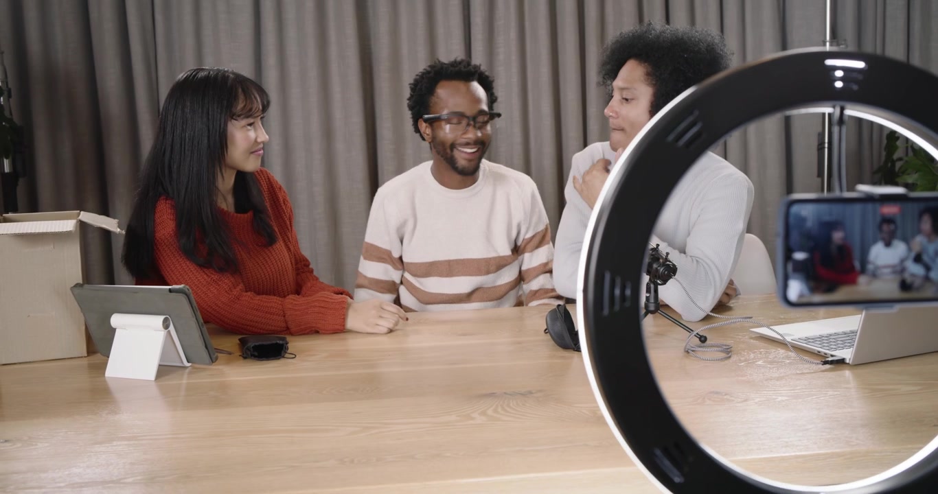 Diverse hosts adjusting to ring light framing phone placing glasses podcasting at wooden table