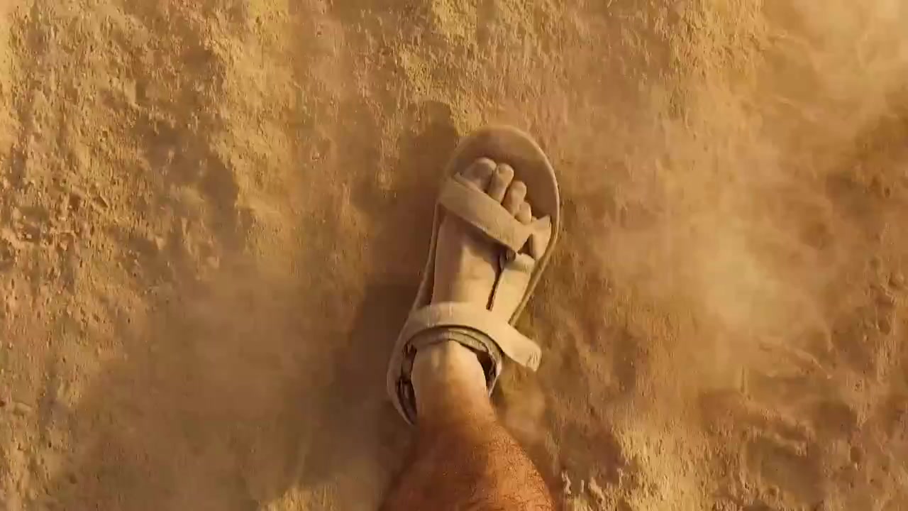 Primo piano in prospettiva in prima persona che guarda i sandali di un uomo che cammina velocemente sulla sabbia in un deserto soleggiato, Israele