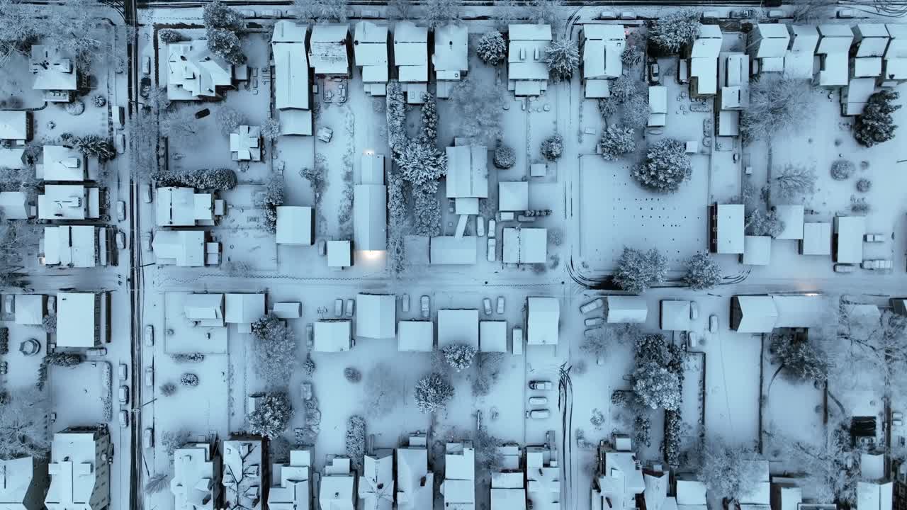 Aerial top down of quiet American residential neighborhood covered in fresh snow. Detached houses line straight street, with snow-covered roofs, trees, sidewalks and parked cars in winter,