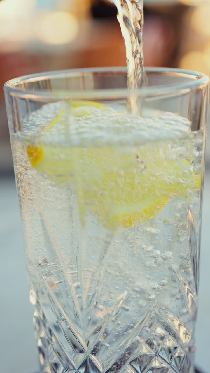 Close up of sparkling water pouring over a slice of lemon in a glass at a terrace. Vertical