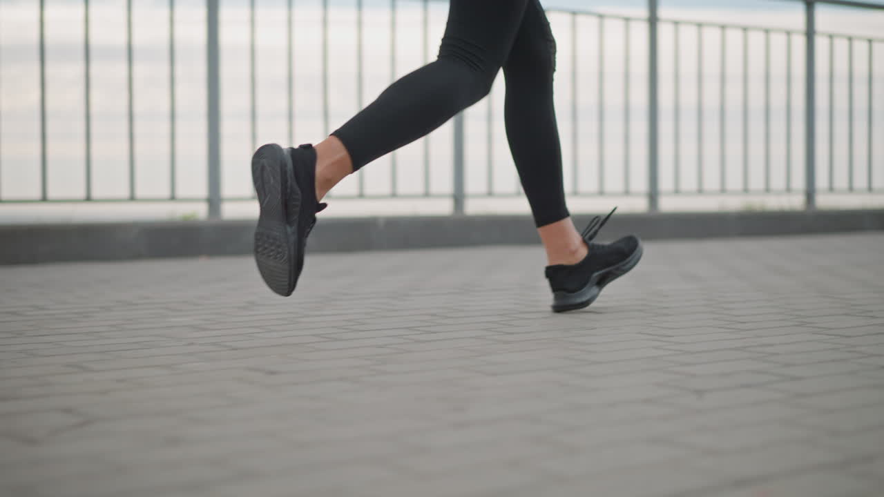 Siyah tayt ve siyah spor ayakkabılarıyla demir çitin yanındaki taş döşeli yolda koşu yapan bir kadının bacaklarının yakın çekimi, açık hava fitness'ında atletik hareketi sergiliyor.