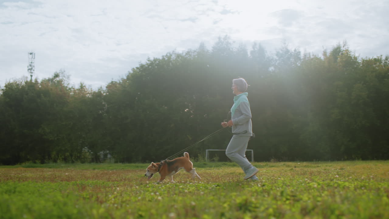 Instructor jogging through football field on sunny day with excited Golden Retriever by side, lush green grass underfoot, background of tall trees, outdoor training session full of energy