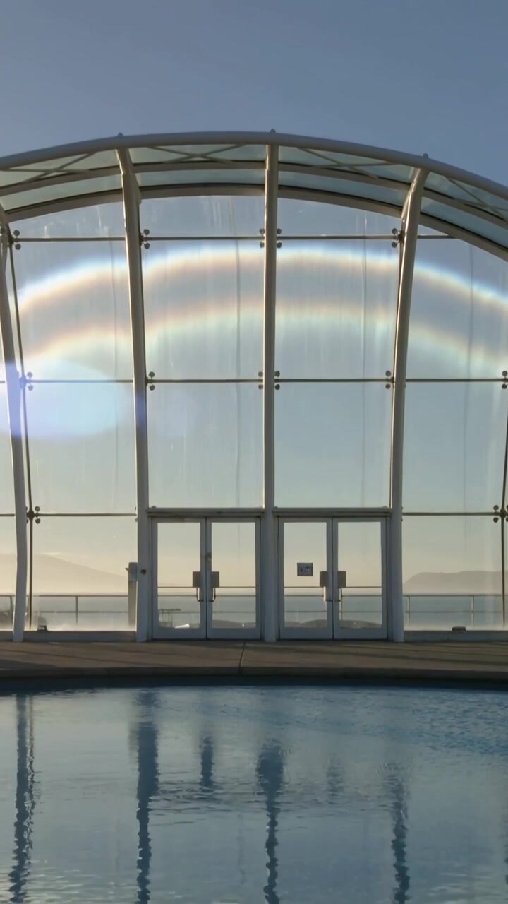 Video vertical: Puertas curvas y relucientes de un pabellón reflejando el arco del sol mientras la brisa riza la piscina en la orilla