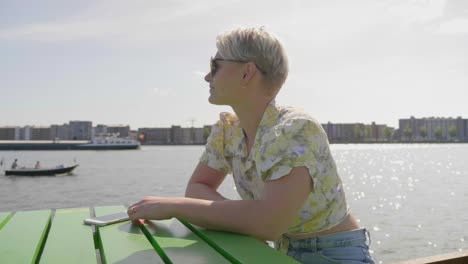 trendy millennial woman watches boats sailing in the river, sitting at a picnic table on the pier