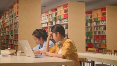 asian woman student with headphones having a headache while typing on a laptop and sitting with her classmate studying on a table in the library