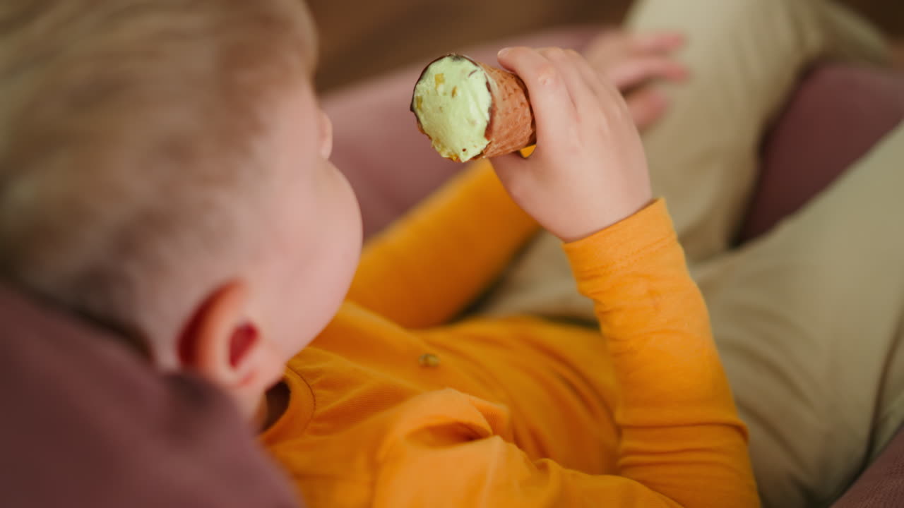 Rear view of young boy in orange shirt holding ice cream cone while sitting barefoot on pink beanbag indoors, enjoying sweet treat with relaxed posture