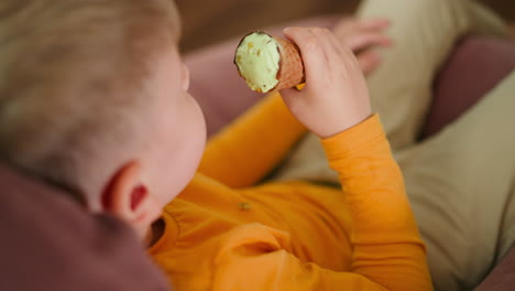 rear view of young boy in orange shirt holding ice cream cone while sitting barefoot on pink beanbag indoors, enjoying sweet treat with relaxed posture
