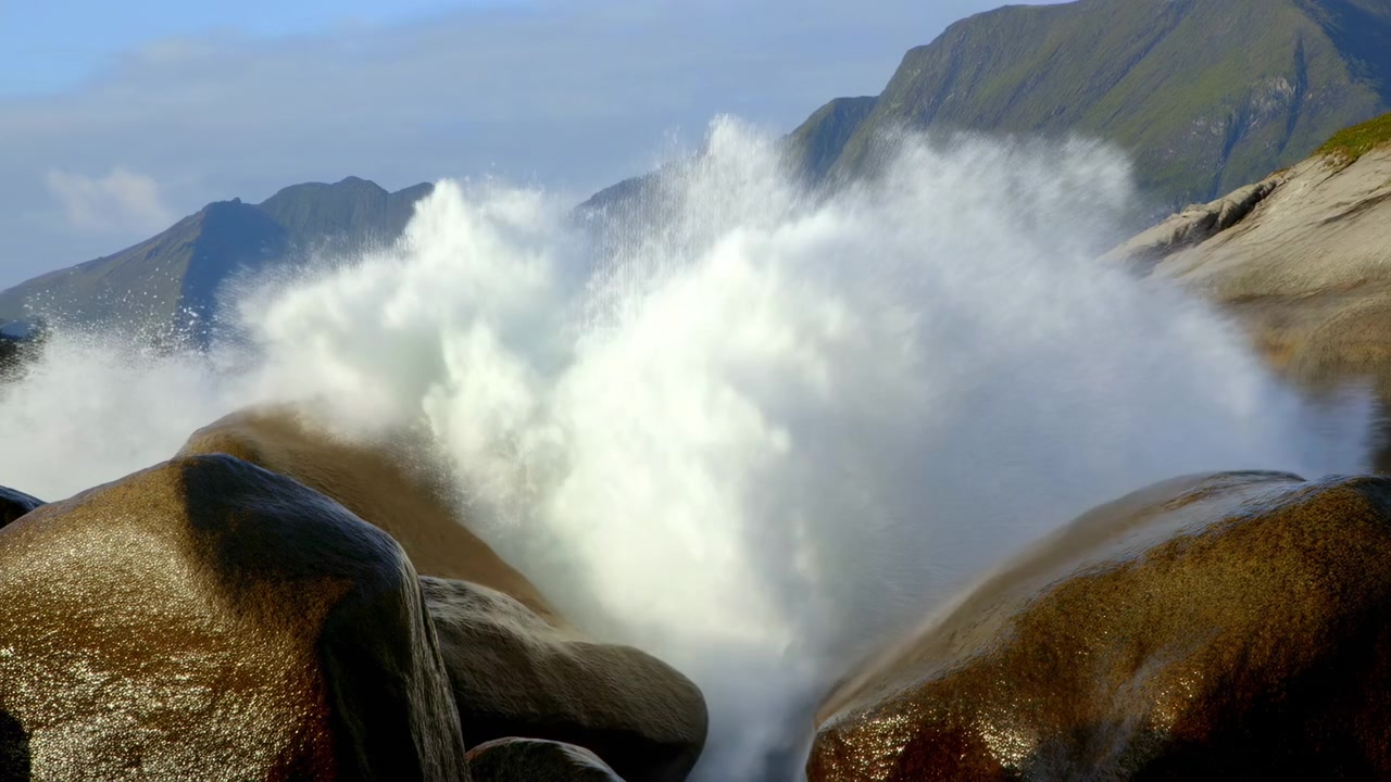 La risacca si infrange tra grandi rocce bagnate su una costa frastagliata, con montagne verdi e un orizzonte lontano sotto una luce diurna soffusa.