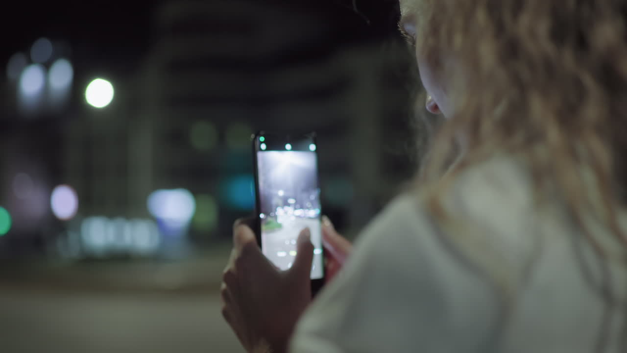 Woman holding smartphone captures illuminated street scene with passing car at night, finger touching screen, soft bokeh lights in urban setting add depth and mood to late evening