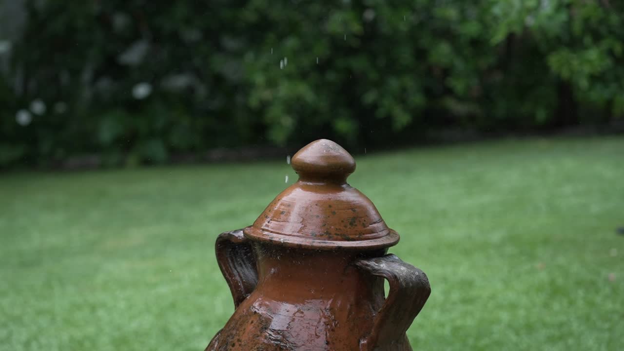 wet terracotta jar with lid under gentle rain in garden with green grass and blurred trees