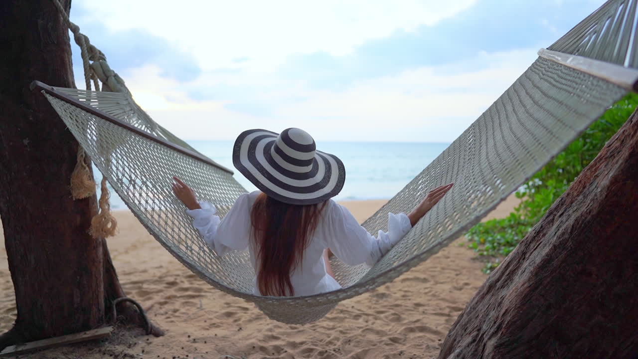 mujer asiática sentada y columpiándose en una hamaca en una playa tropical en el paisaje marino de tailandia en la vista trasera de cámara lenta de fondo